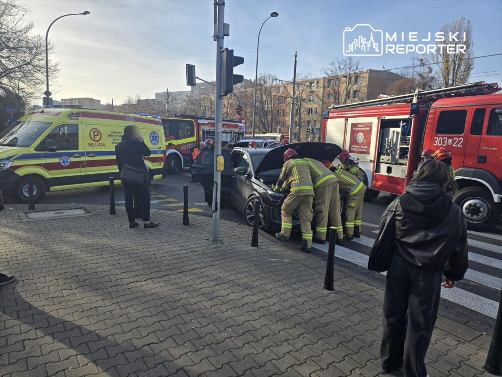 Grupa strażaków sprawdzających silnik samochodu na skrzyżowaniu, obok stoi karetka pogotowia.