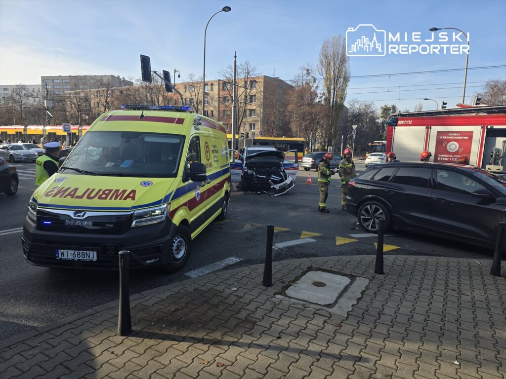 Ambulans stoi na skrzyżowaniu, a strażacy zabezpieczają miejsce wypadku z udziałem dwóch samochodów.
