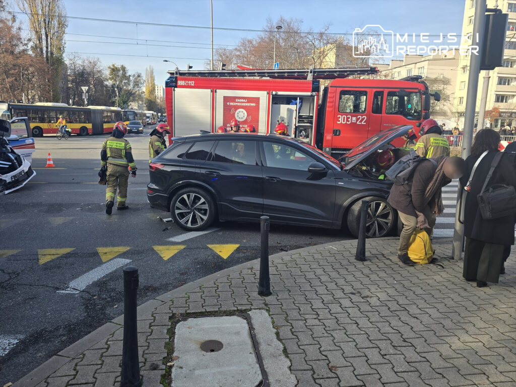 Wypadek drogowy z udziałem czarnego samochodu osobowego na skrzyżowaniu, w tle strażacy oraz pojazd strażacki.