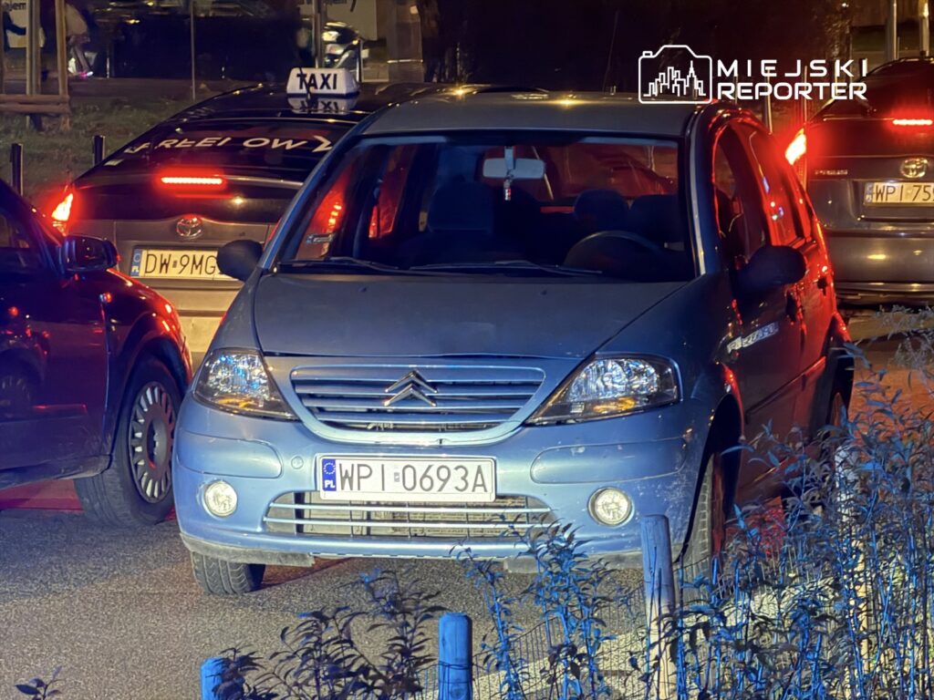 Niebieski samochód osobowy stoi w korku obok taksówki na oświetlonej ulicy. W tle widać inne pojazdy.