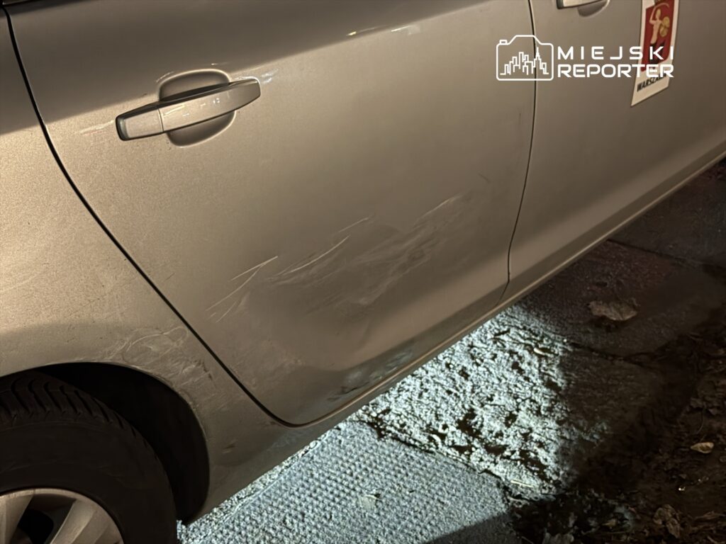 Zarysowany bok srebrnego samochodu zaparkowanego na oświetlonym chodniku.