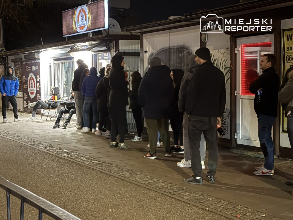 Grupa osób czeka w kolejce przed budynkiem z oświetlonymi witrynami, w nocy, na ulicy.