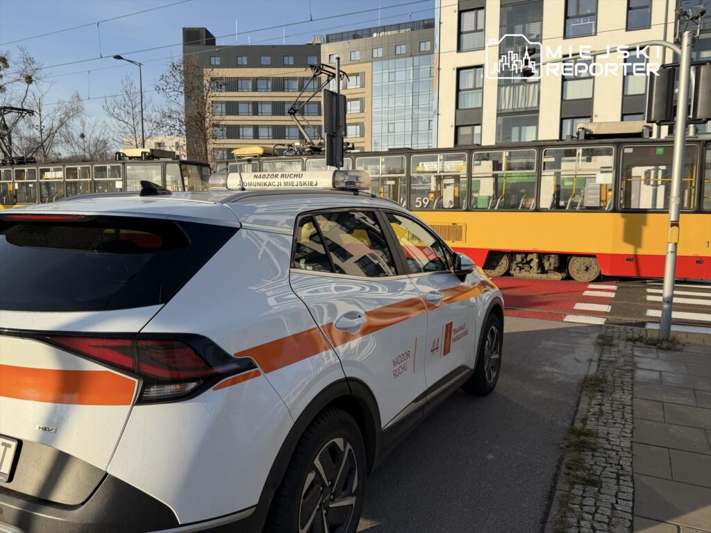 Samochód z oznaczeniem "Nadzór Ruchu" stoi na skrzyżowaniu obok tramwaju, w tle nowoczesne budynki.
