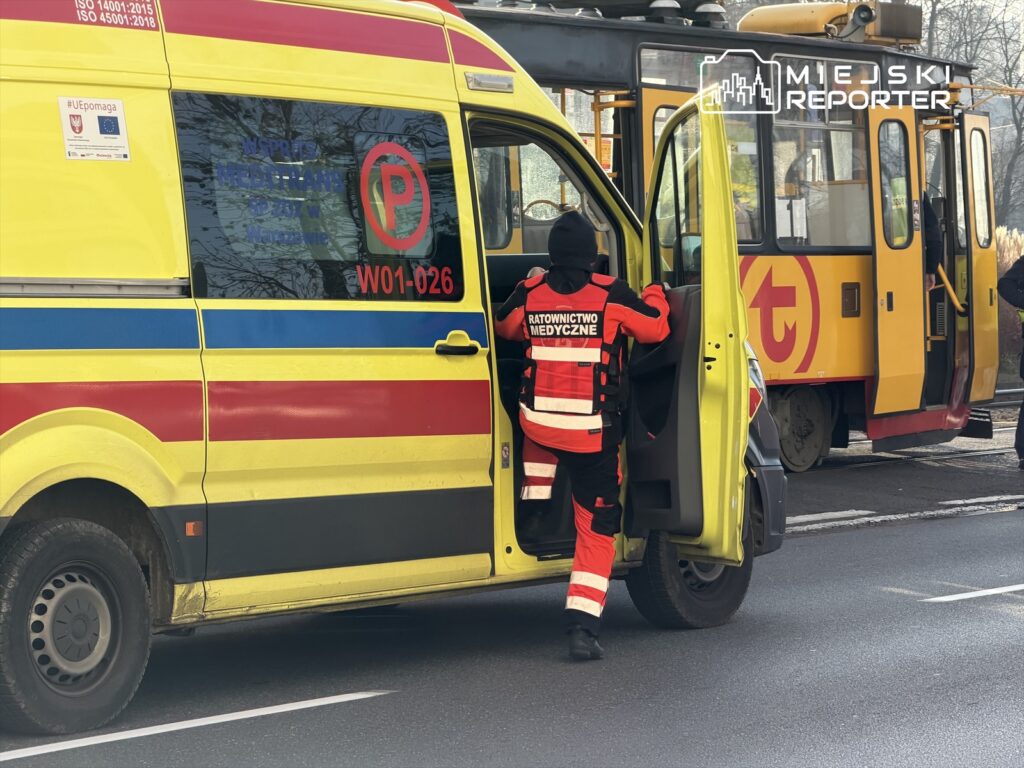Ratownik medyczny w czerwono-czarnym uniformie wchodzi do żółtej karetki stojącej obok tramwaju na ulicy.