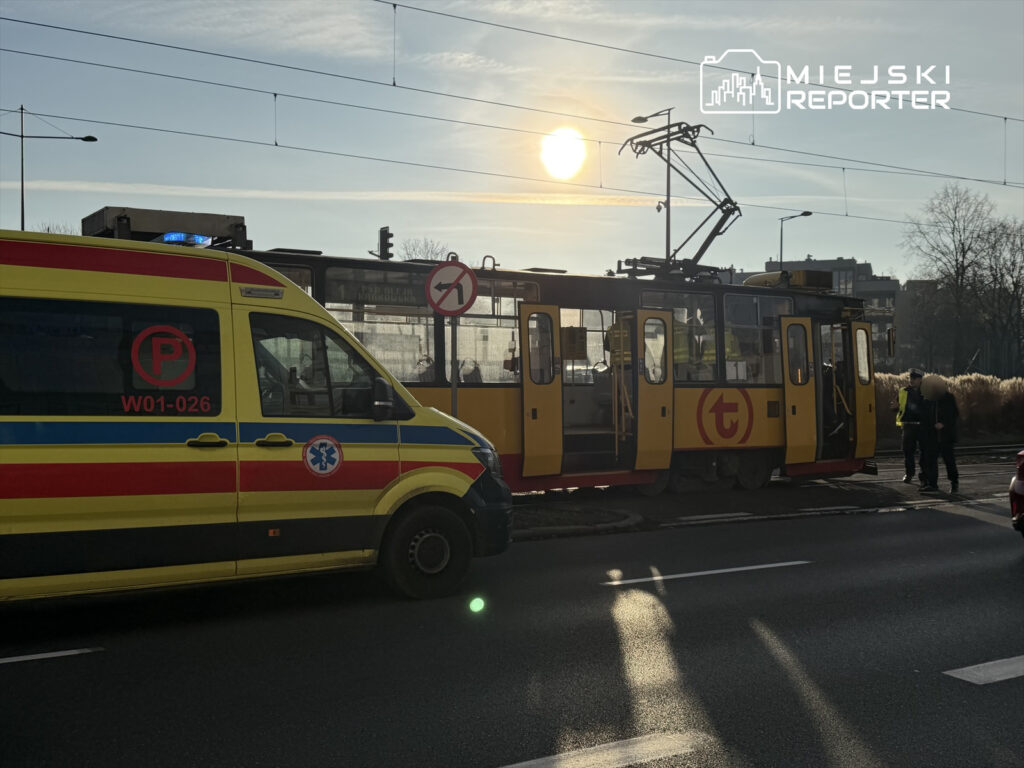 Karetka pogotowia z włączonymi sygnałami świetlnymi stoi obok tramwaju na ulicy, w tle widać wschodzące słońce.