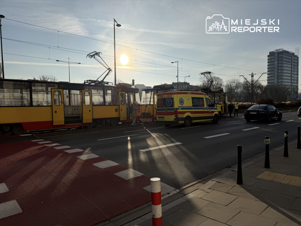 Tramwaj w żółto-czerwonych barwach zatrzymany na torach, obok stoi karetka pogotowia, a w tle widać nowoczesny biurowiec.