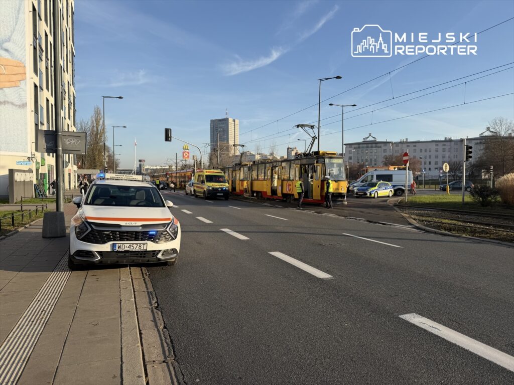 Karetkę i radiowóz policji przy torach tramwajowych, obok zatrzymane tramwaje, w tle nowoczesne budynki.