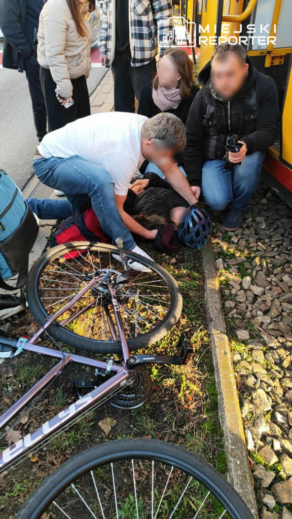 Grupa osób otacza leżącego na ziemi rowerzystę, a jeden mężczyzna udziela mu pomocy, obok leży przewrócony rower.