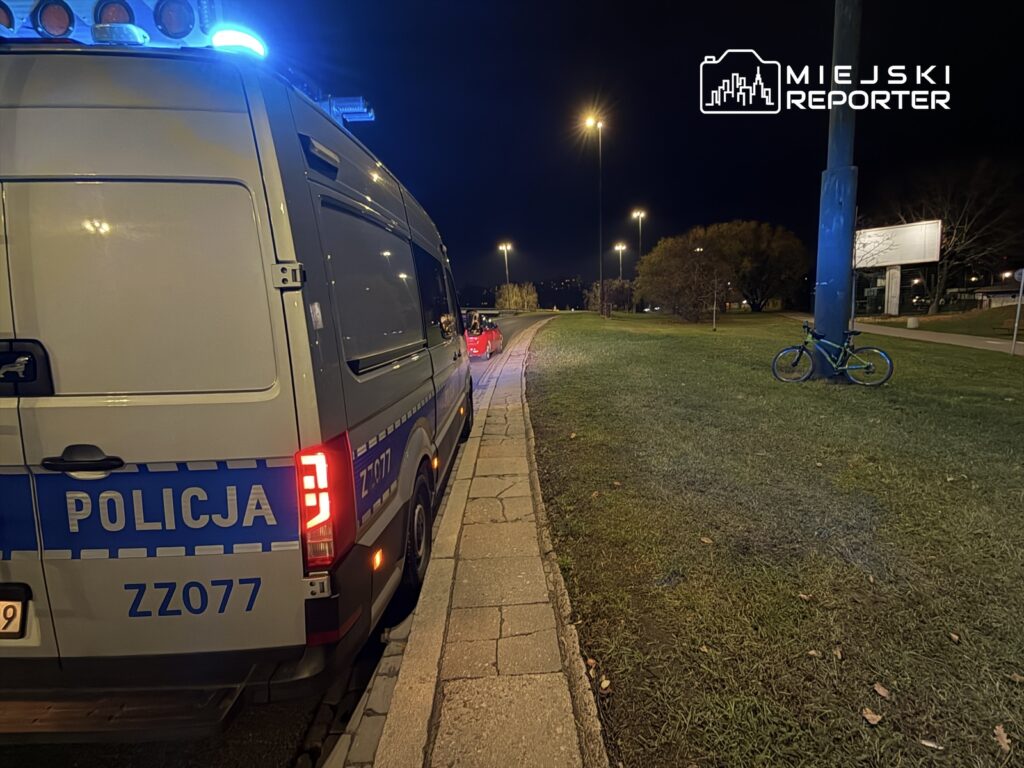 Policyjny van z włączonymi sygnałami świetlnymi stoi na poboczu drogi nocą, obok znajduje się zielony rower.