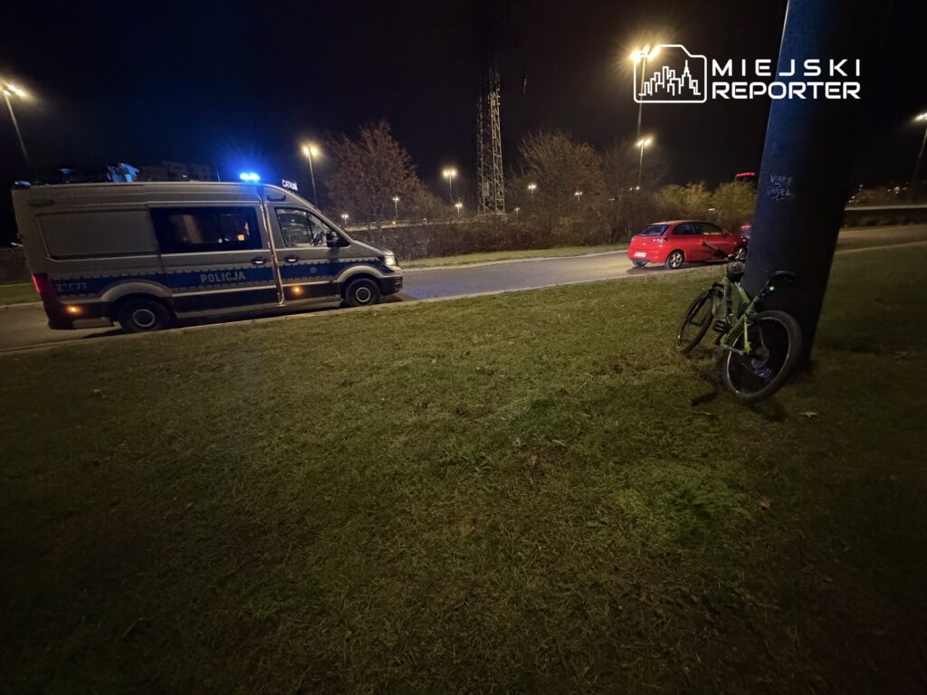 Policyjny van z włączonymi sygnalizatorami stoi na trawie obok drogi, a zielony rower oparty jest o latarnię.