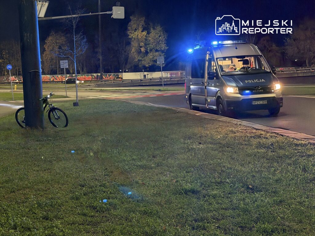 Policyjny furgon z włączonymi sygnałami świetlnymi stoi na poboczu, obok zaparkowany rower.
