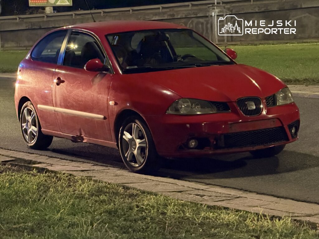 Czerwony samochód osobowy zaparkowany na poboczu drogi nocą, z widocznymi zarysowaniami na karoserii.