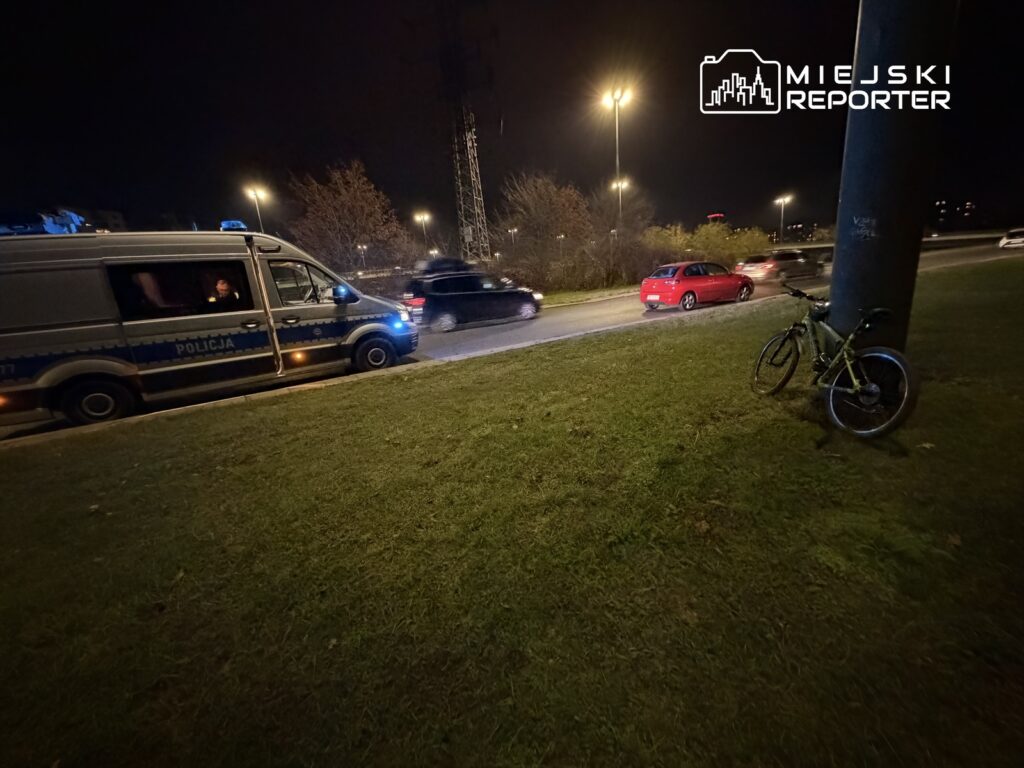 Policyjny furgon zatrzymany przy drodze, obok stoi zielony rower, a w tle poruszające się samochody.