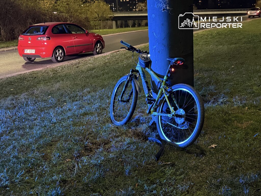 Zielony rower z podświetleniem stoi obok słupa, a w tle widoczny jest czerwony samochód zaparkowany na poboczu.