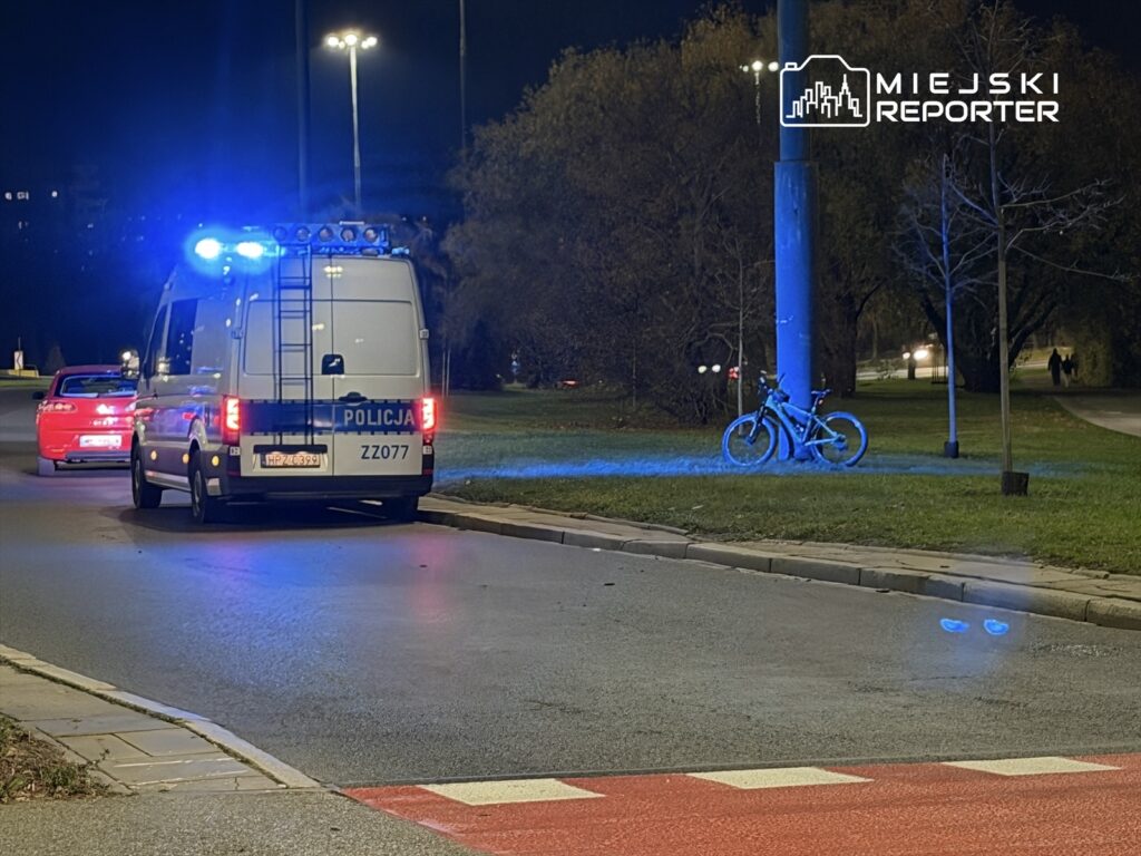 Policyjny radiowóz z włączonymi sygnałami świetlnymi stoi na ulicy obok zaparkowanego czerwonego samochodu, a w tle widać rowery.
