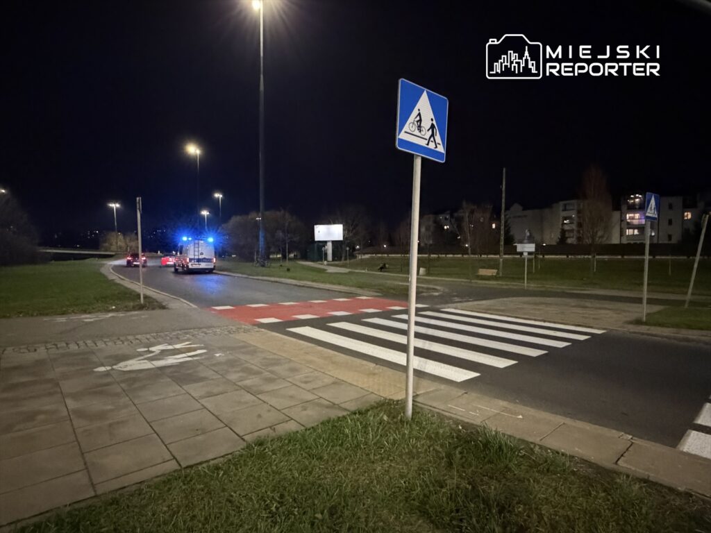 Nocna scena na skrzyżowaniu, gdzie radiowóz z włączonymi sygnałami świetlnymi zatrzymuje się przed przejściem dla pieszych.