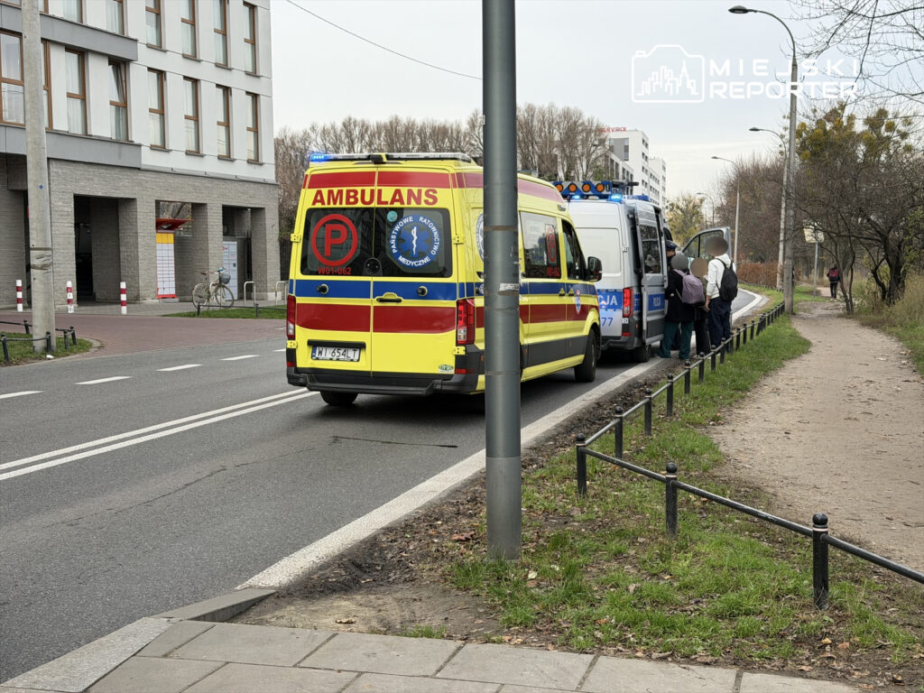 Ambulans zatrzymany przy drodze, a obok grupa osób rozmawia z policjantami w miejskim otoczeniu.