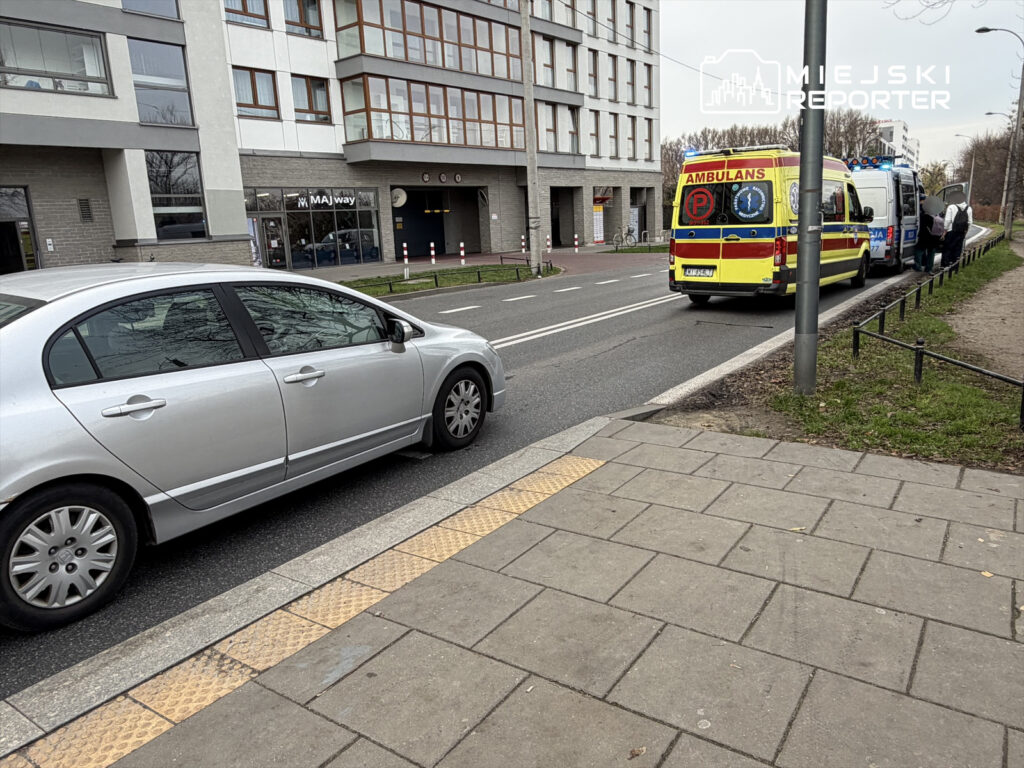 Ambulans zatrzymany przy poboczu ulicy, obok stoi kilka osób, a w tle nowoczesny budynek z dużymi oknami.