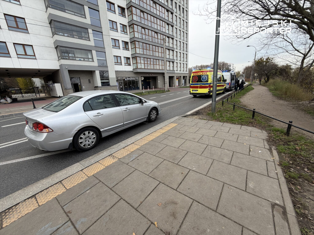 Srebrny samochód osobowy zatrzymany na ulicy obok karetki pogotowia, w tle nowoczesny biurowiec.