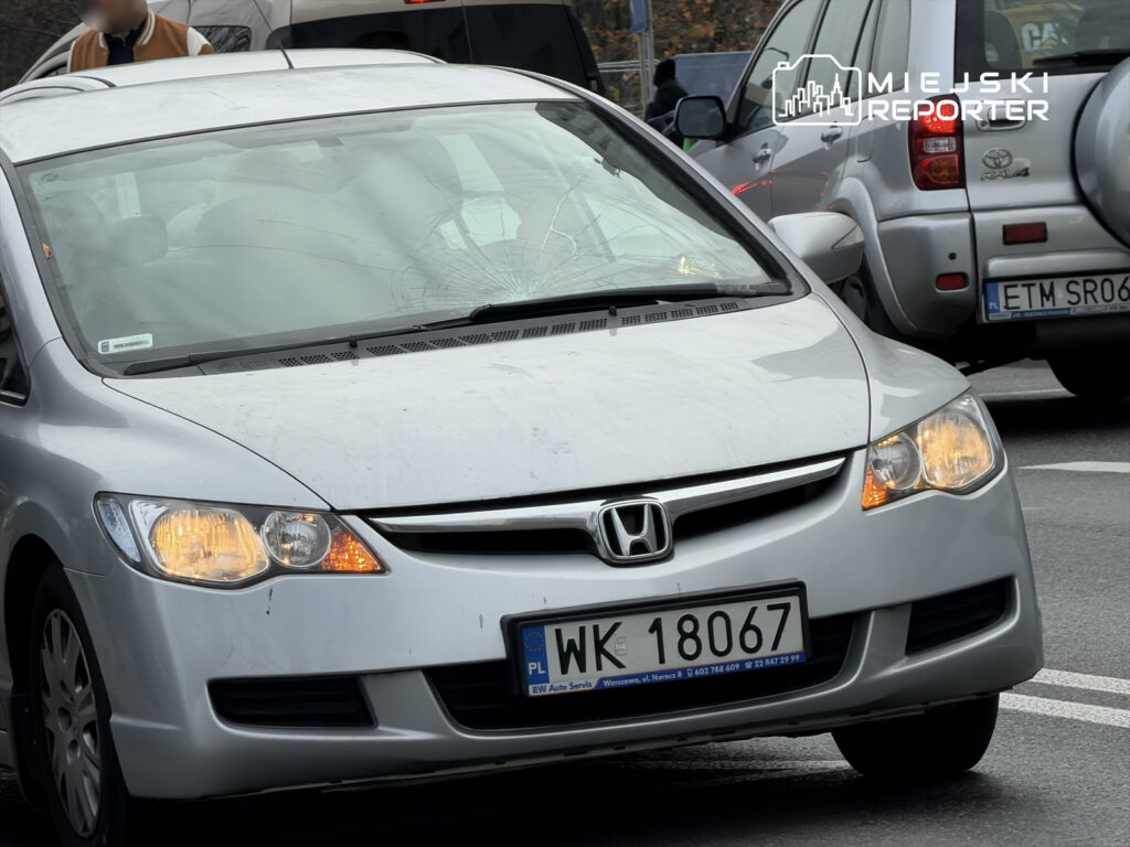 Srebrny samochód osobowy marki Honda z uszkodzoną szybą przednią porusza się w ruchu miejskim.