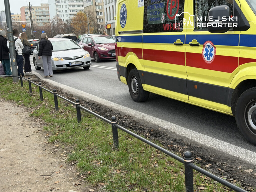Grupa osób stoi na chodniku obok ruchliwej ulicy, podczas gdy karetka pogotowia zatrzymuje się na jezdni.