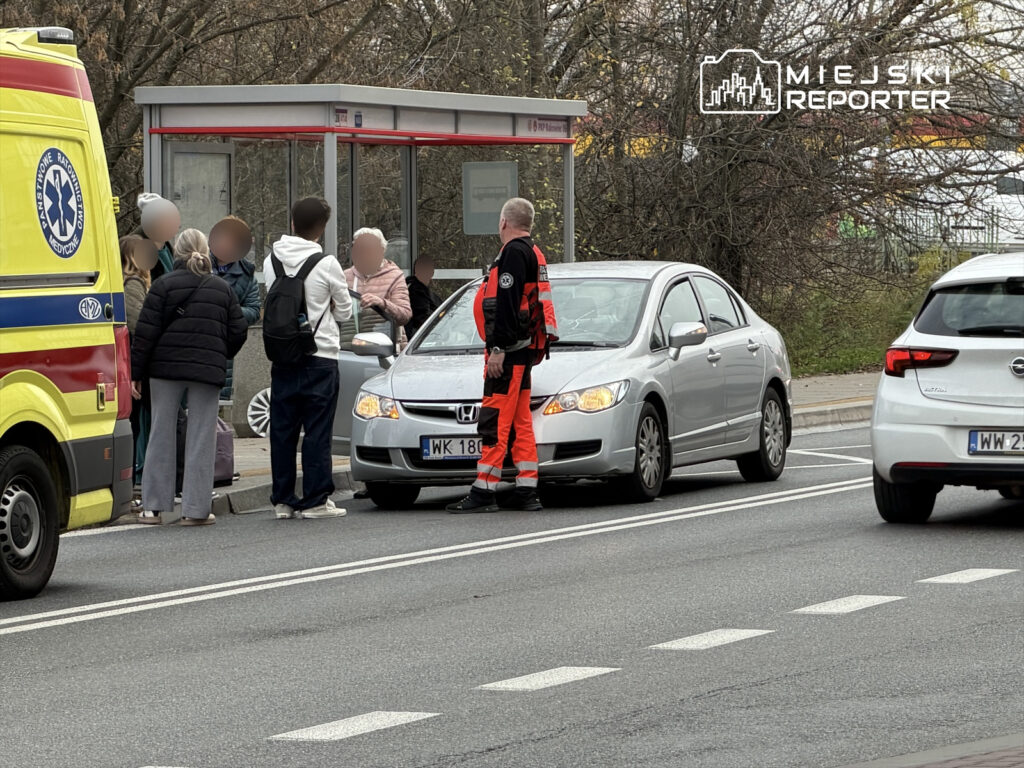 Grupa ludzi stojąca przy przystanku autobusowym, rozmawia z ratownikiem w czerwonym uniformie obok samochodu osobowego.