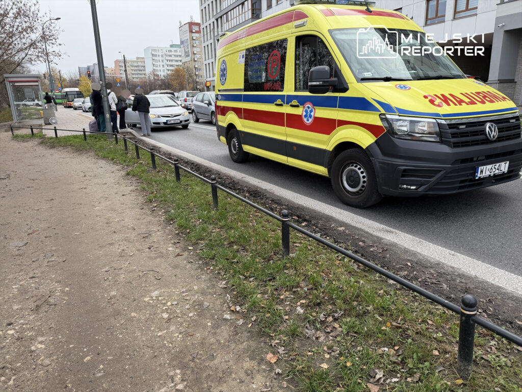 Grupa osób czeka obok przystanku na ulicy, obok stoi karetka pogotowia w intensywnych barwach.