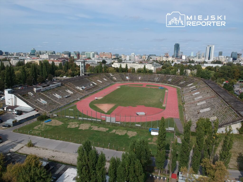 Stadion z bieżnią, pustymi trybunami i zielonym boiskiem, otoczony nowoczesnymi budynkami w tle.