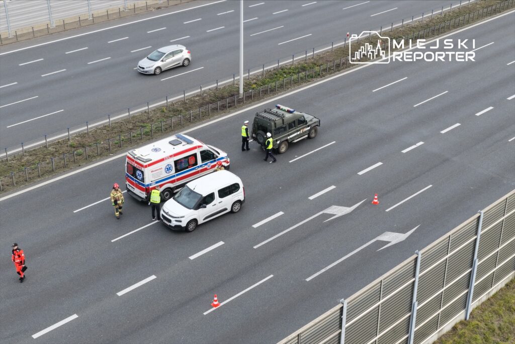Na jezdni zatrzymane pojazdy: karetka, samochód osobowy i radiowóz; w tle strażacy i policjanci prowadzący rozmowy.