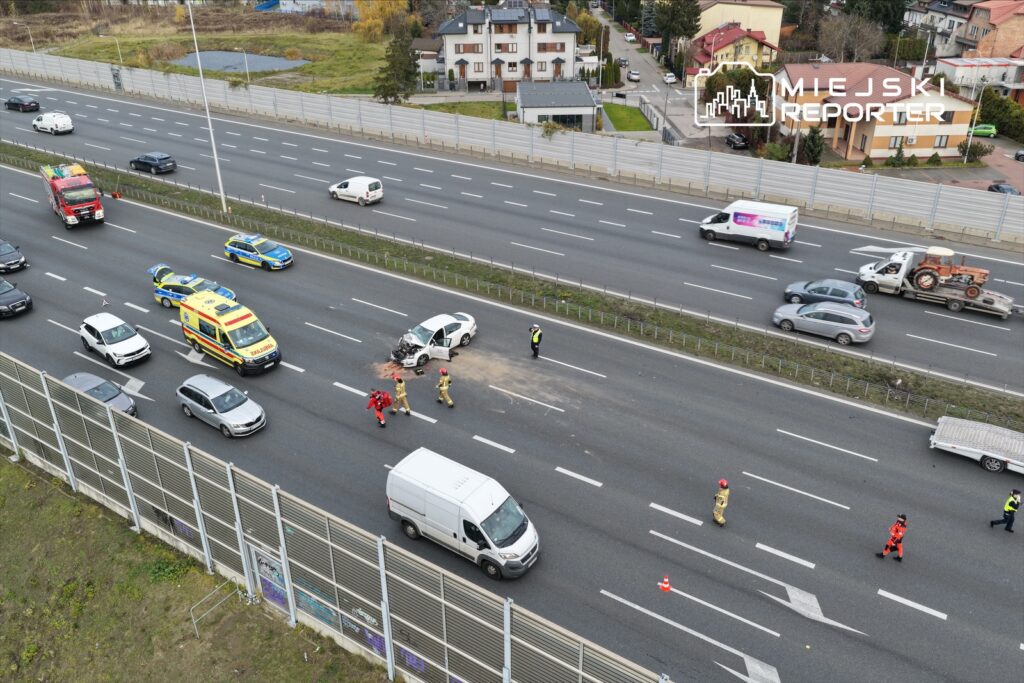 Na drodze widoczny jest wypadek samochodowy, w którym uczestniczy uszkodzony pojazd oraz ekipy ratunkowe.