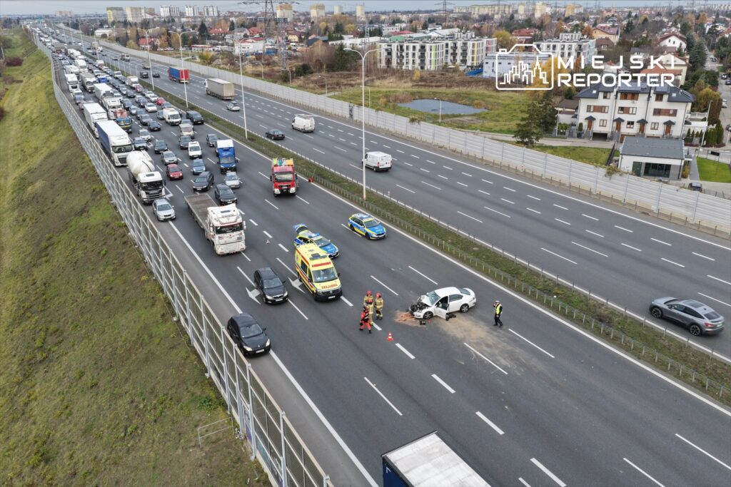 Na autostradzie zatrzymane pojazdy, w tym białe auto z uszkodzeniem, a obok interweniują strażacy i policja.