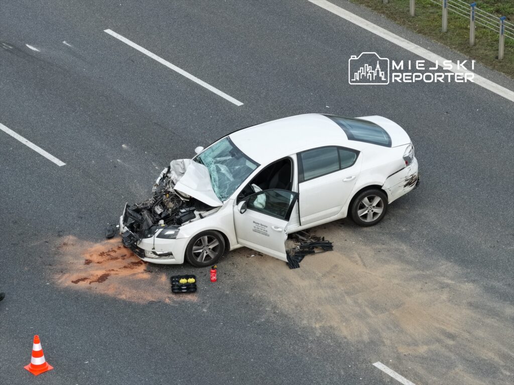 Biały samochód osobowy z uszkodzonym przodem stoi na jezdni, obok widoczne plamy płynów i narzędzia.