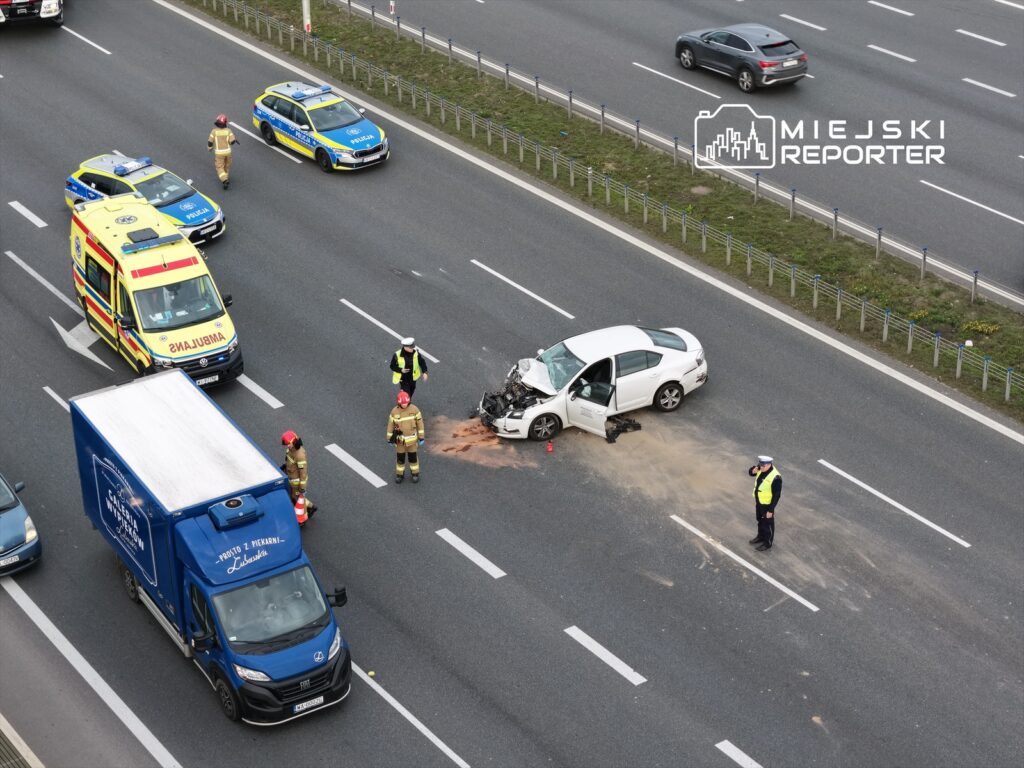 Na drodze leży uszkodzony biały samochód, obok niego pracują strażacy i policjant, a w tle widać ambulans.