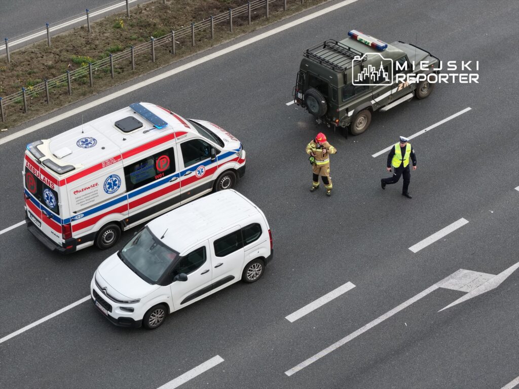 Na drodze zatrzymane pojazdy: karetka pogotowia, samochód osobowy i policyjny, obok biegający strażak oraz policjant.
