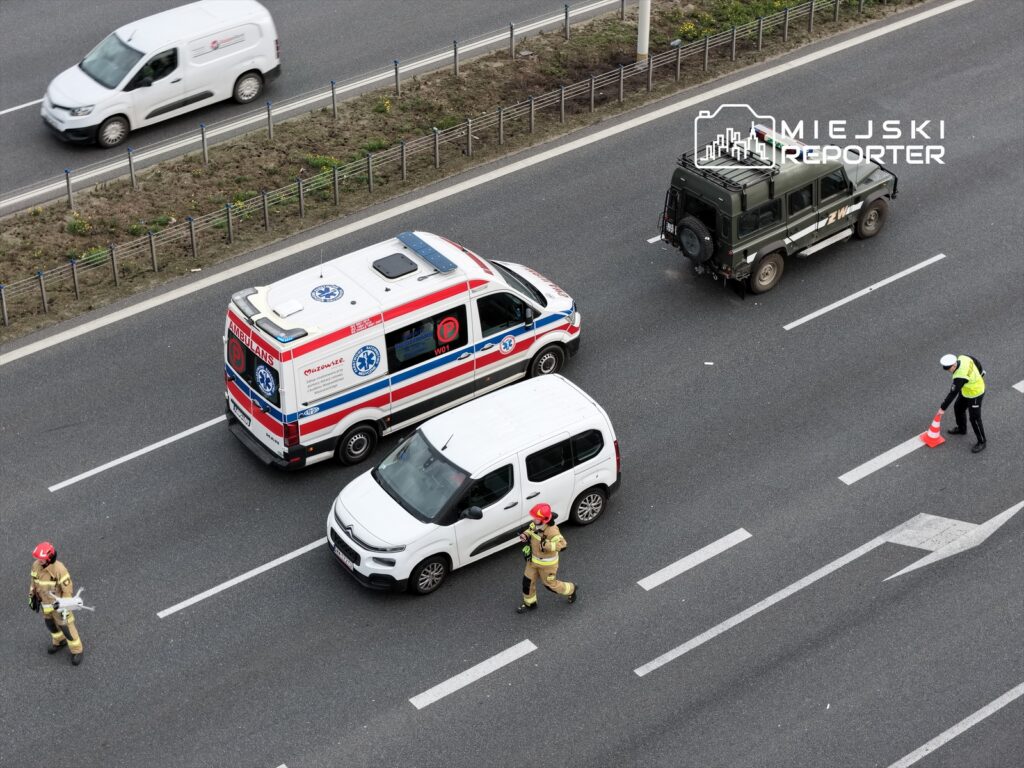 Ratownicy medyczni i policjant kierują ruchem na drodze, obok stoi karetka i pojazd służbowy.