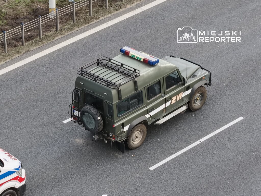 Samochód terenowy w kolorze zielonym z oznaczeniem "ZWI" stoi na drodze, obok widoczny pojazd służb ratunkowych.
