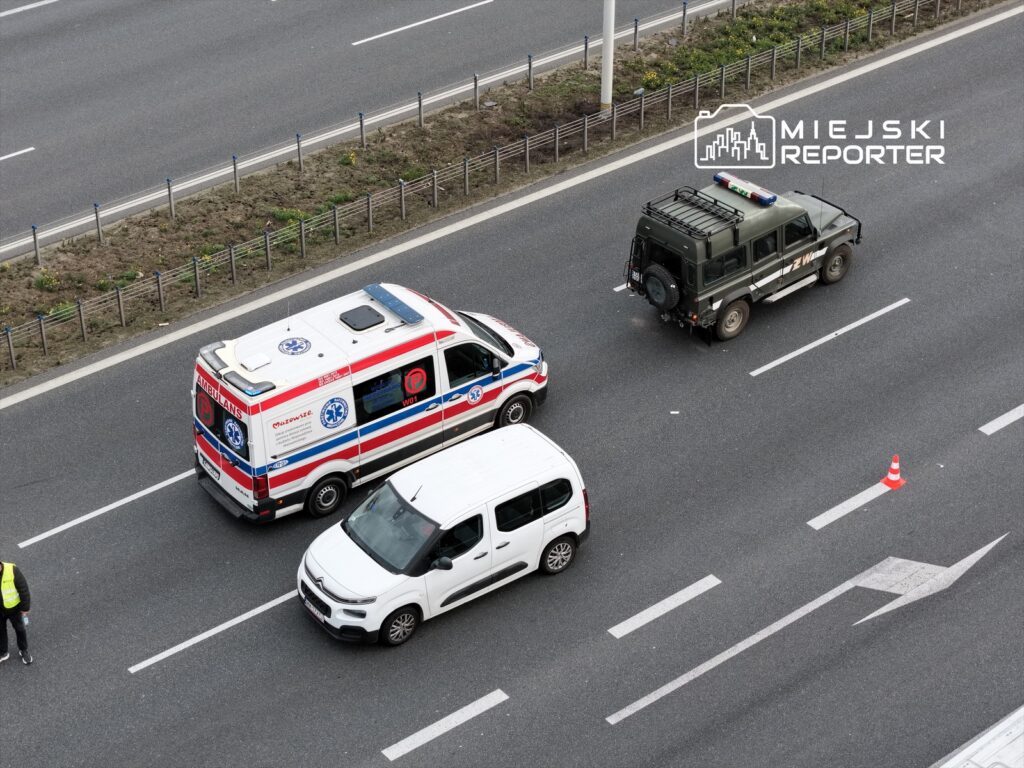 Karetka pogotowia i pojazd policyjny zatrzymane na drodze, obok biały samochód osobowy oraz człowiek w kamizelce odblaskowej.