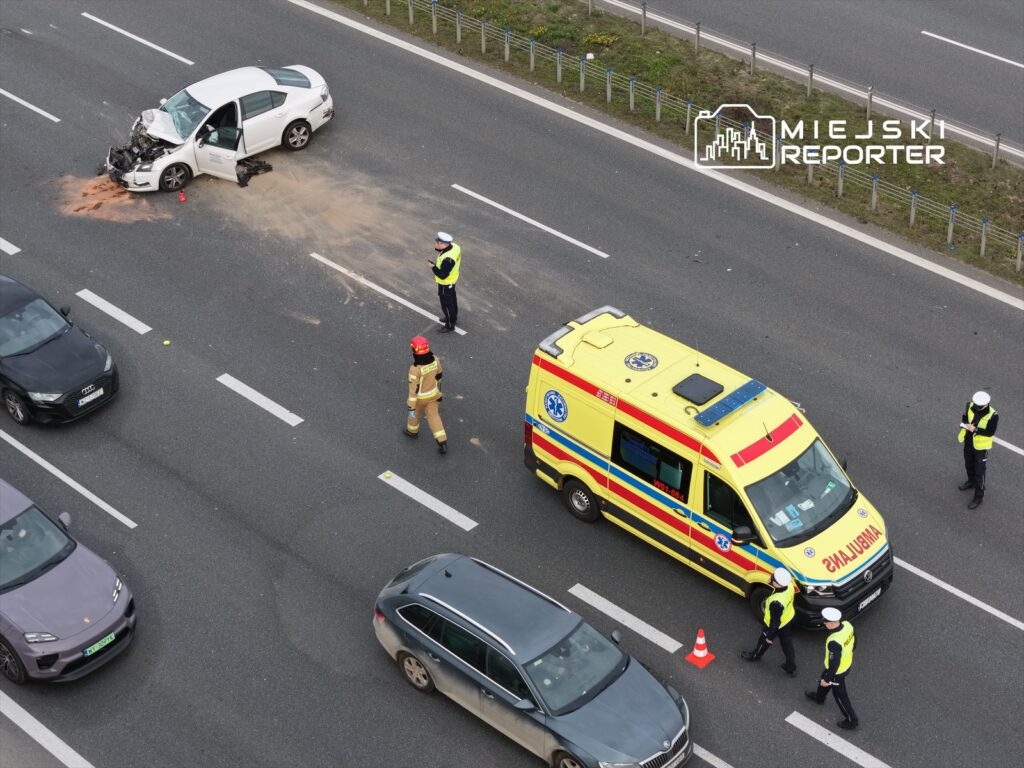 Wypadek drogowy z uszkodzonym białym samochodem na jezdni, obok którego pracują służby ratunkowe i policja.