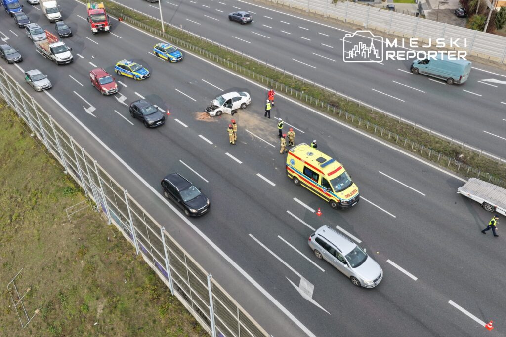 Na drodze widoczny wypadek samochodowy, wokół rozstawione pachołki, a ratownicy medyczni i strażacy udzielają pomocy.
