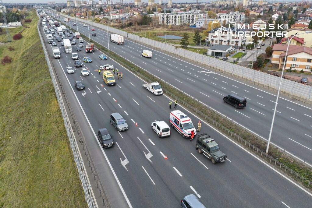 Na drodze szybkiego ruchu grupa ratowników i policjantów zabezpiecza miejsce wypadku, obok stoją pojazdy służb ratunkowych.