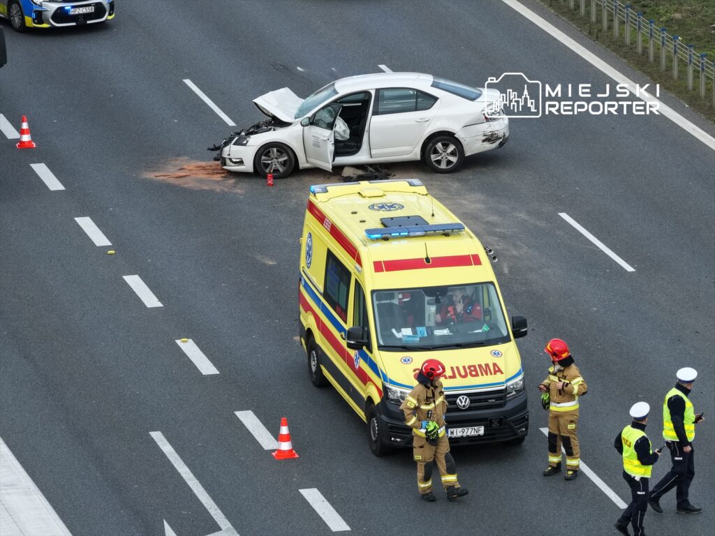 W białym samochodzie z otwartą maską leży plama płynu, obok stoi karetka i strażacy w hełmach.