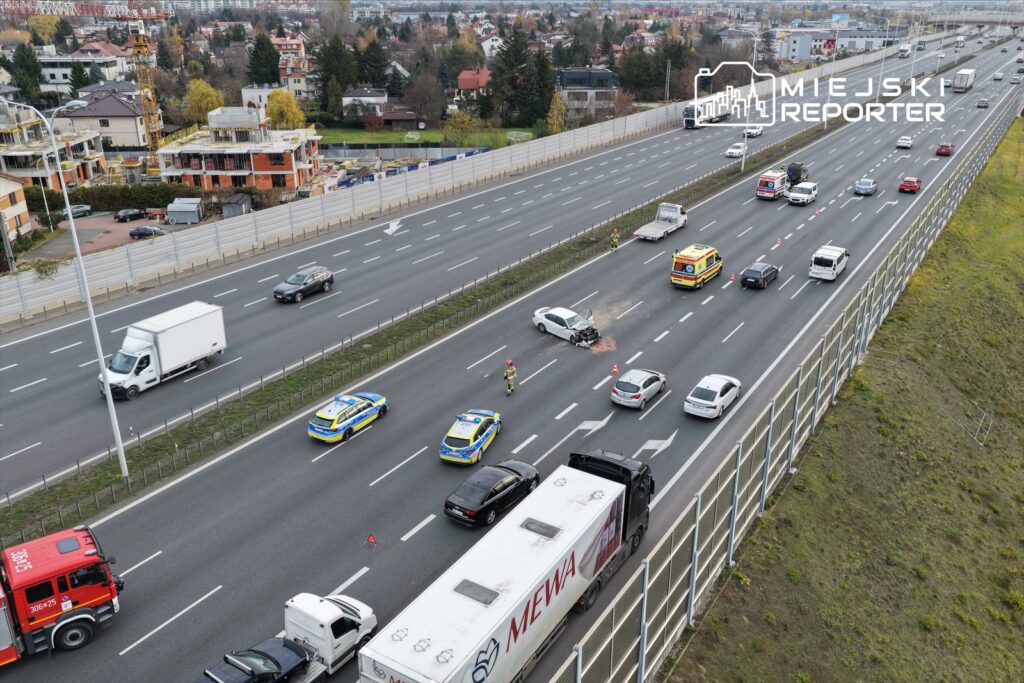 Na drodze krajowej widać samochód osobowy z uszkodzoną maską, a obok niego pracują strażacy oraz policja.