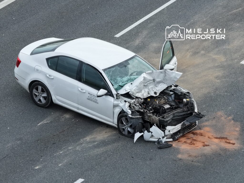 Biały samochód osobowy z uszkodzoną maską i wybitym szybą stoi na drodze, obok widoczne ślady po wypadku.