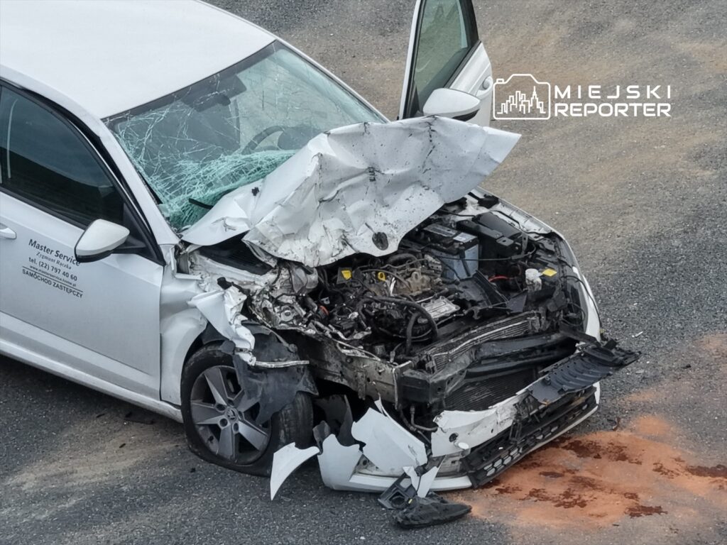 Uszkodzony samochód osobowy z wgniecionym przodem stoi na żwirowym parkingu, z pękniętą szybą i wyciekającymi płynami.