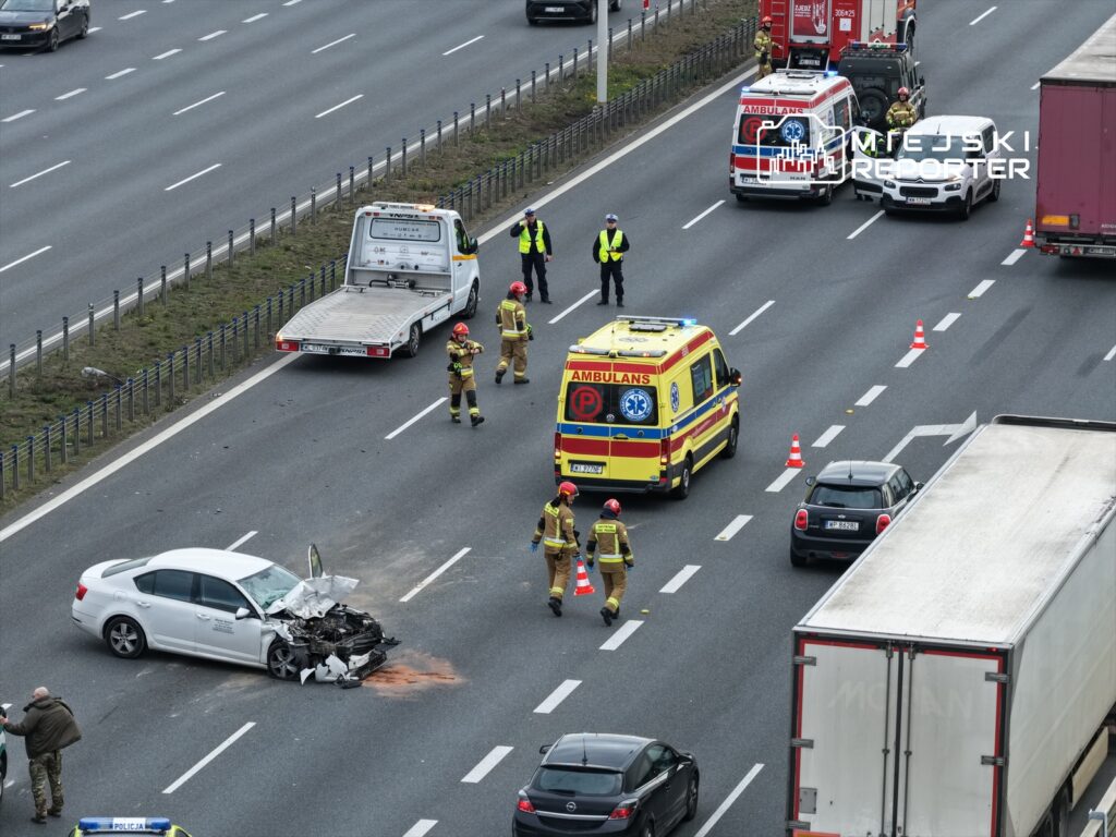 Na drodze stoi uszkodzony biały samochód osobowy, obok pracują strażacy i ratownicy medyczni. W tle widać pojazdy oraz policyjny radiowóz.