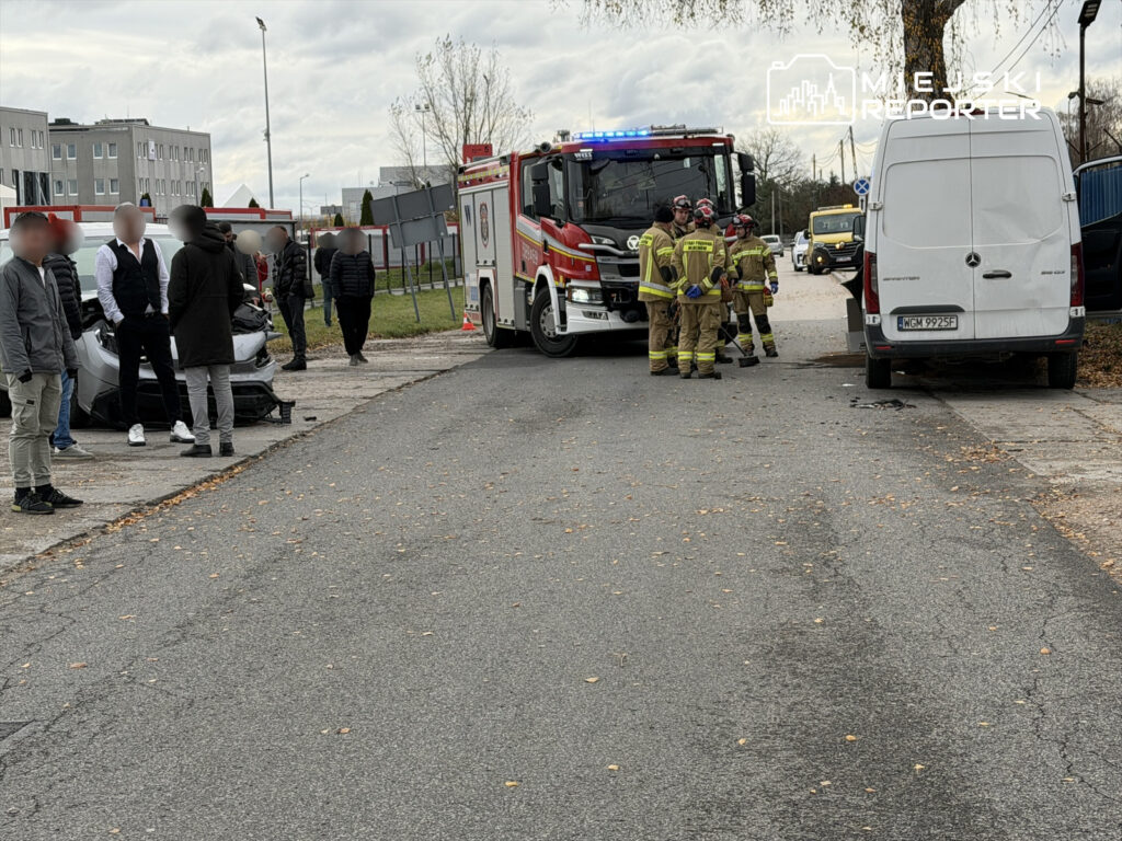 Grupa strażaków w uniformach omawia sytuację przy uszkodzonym samochodzie, obok stoi kilka osób.