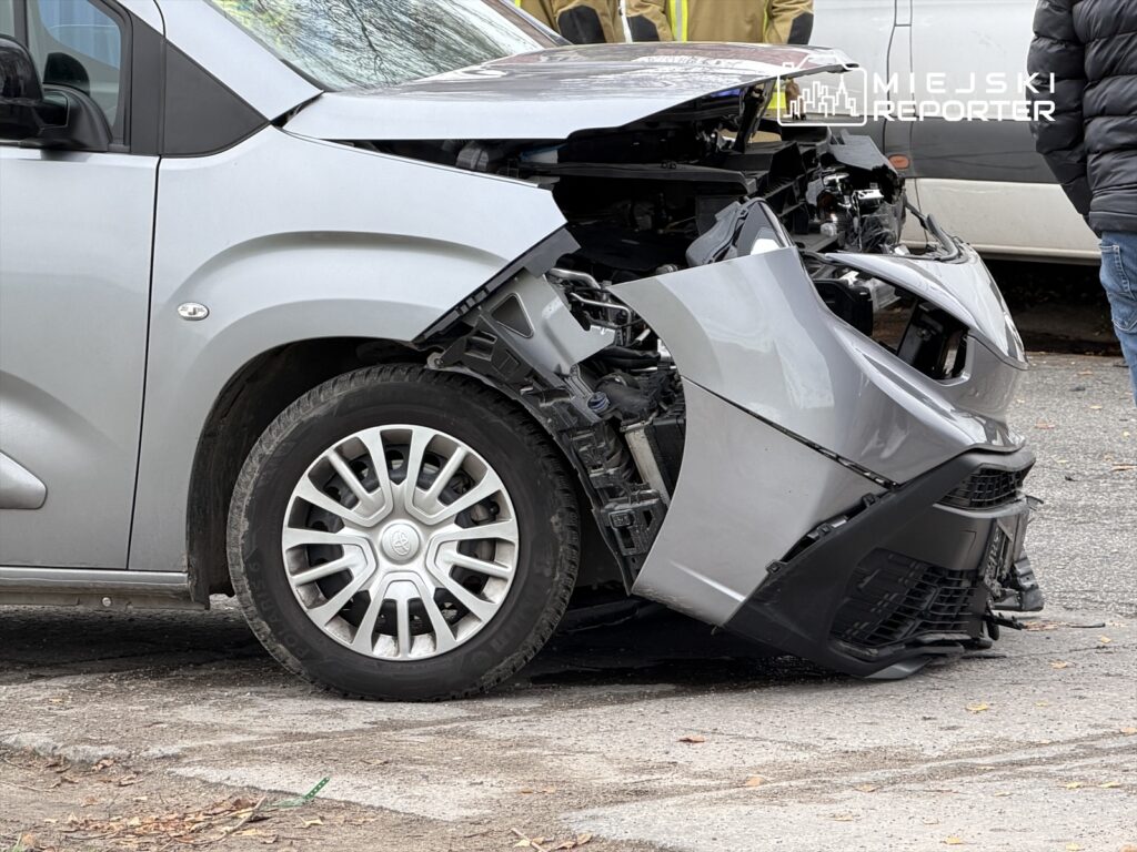 Rozbity samochód osobowy z uszkodzoną przednią częścią stoi na ulicy, obok dwóch osób w odzieży roboczej.