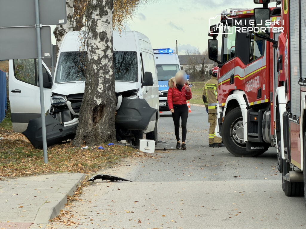 Biały furgon uderzony w drzewo, obok kobieta w czerwonej kurtce rozmawiająca z ratownikami, w tle pojazdy straży pożarnej.