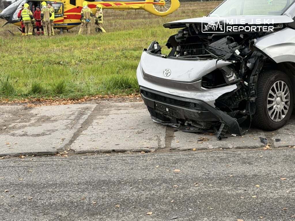 Uszkodzony samochód osobowy stoi na drodze, obok lądowisko dla helikoptera ratunkowego oraz grupa ratowników.