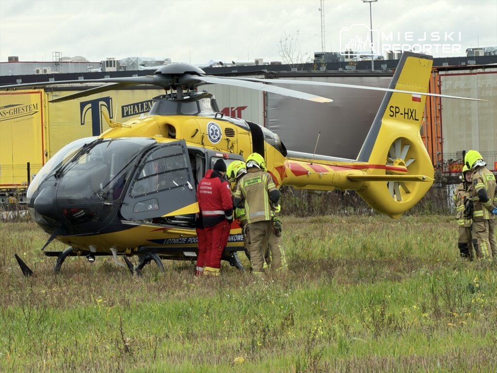 Ratownicy w odzieży ochronnej podchodzą do śmigłowca ratunkowego na zielonej łące w miejskim otoczeniu.